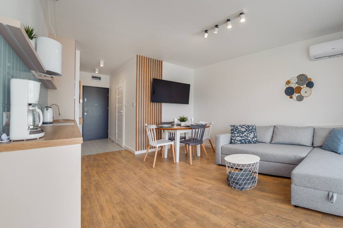 Una sala de estar moderna con un sofá gris, una mesa de centro y una zona de comedor con mesa y sillas. Hay un televisor montado en la pared, un equipo de aire acondicionado y un adorno decorativo.