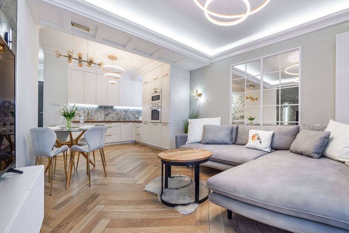 Una sala de estar moderna con un sofá gris, una mesa de comedor con sillas y una cocina con armarios blancos y un salpicadero de mármol.