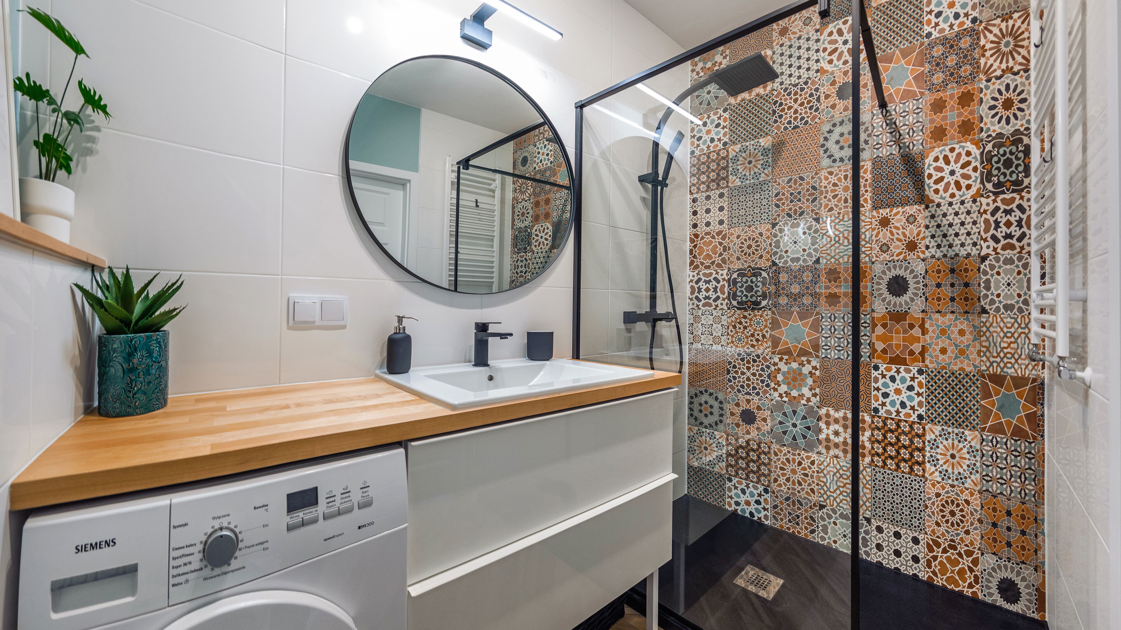 Ein modernes Badezimmer mit einer Holzausziehtheke, einem weißen Waschbecken, einem runden Spiegel, einer integrierten Waschmaschine und einer Dusche mit einer Wand aus gemusterten Fliesen.