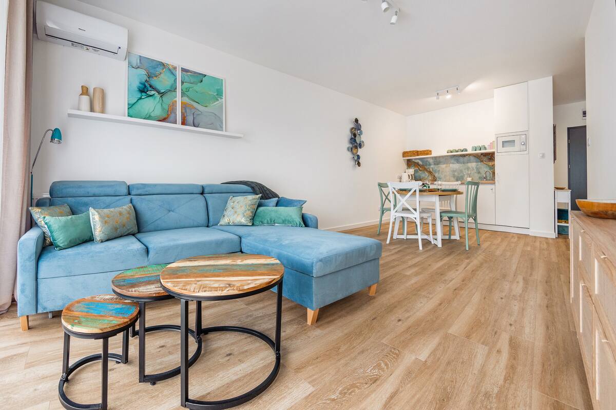 Una sala de estar moderna con un sofá azul, dos mesas redondas y una zona de cocina con una mesa de comedor y sillas.