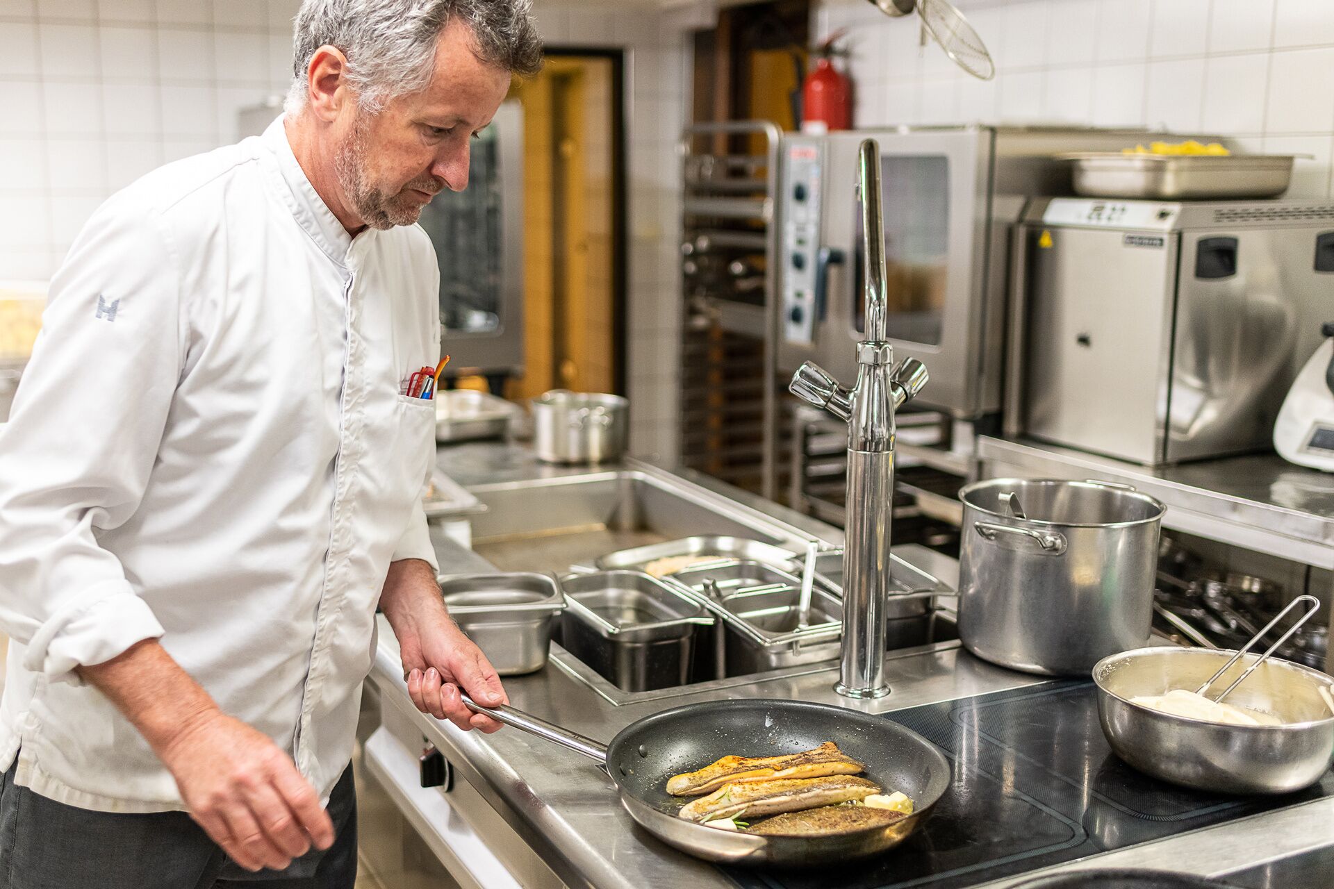 Un chef cocinando salchichas en una cocina profesional con electrodomésticos y utensilios de acero inoxidable.