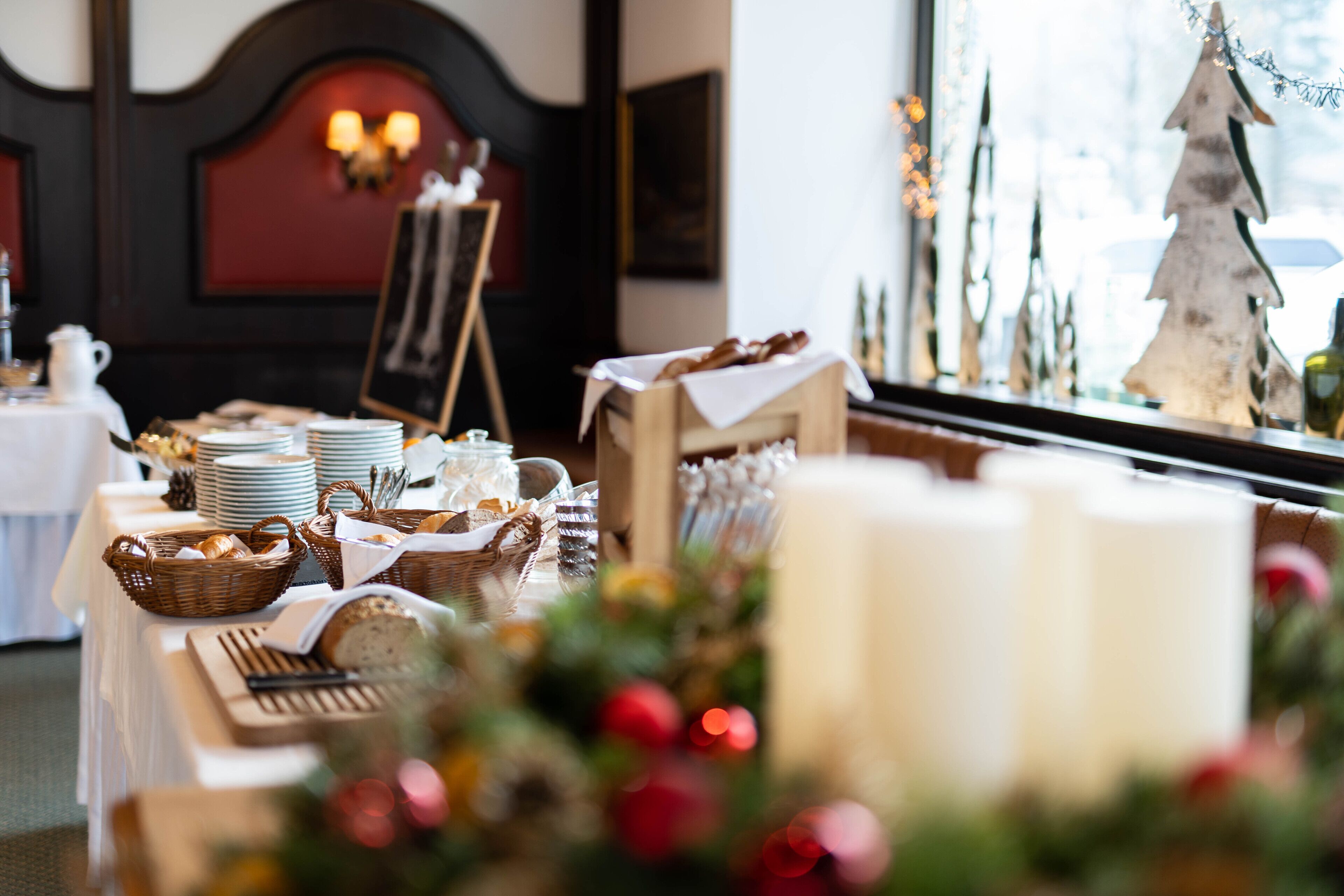Una mesa de comedor dispuesta con platos, cestas y velas, con un árbol de Navidad y adornos al fondo.