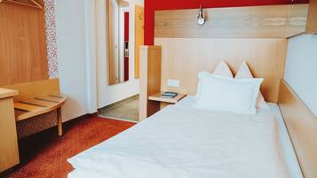 A hotel room with a bed, a bedside table, a nightstand, a wooden headboard, a hanging coat rack, and a patterned curtain.