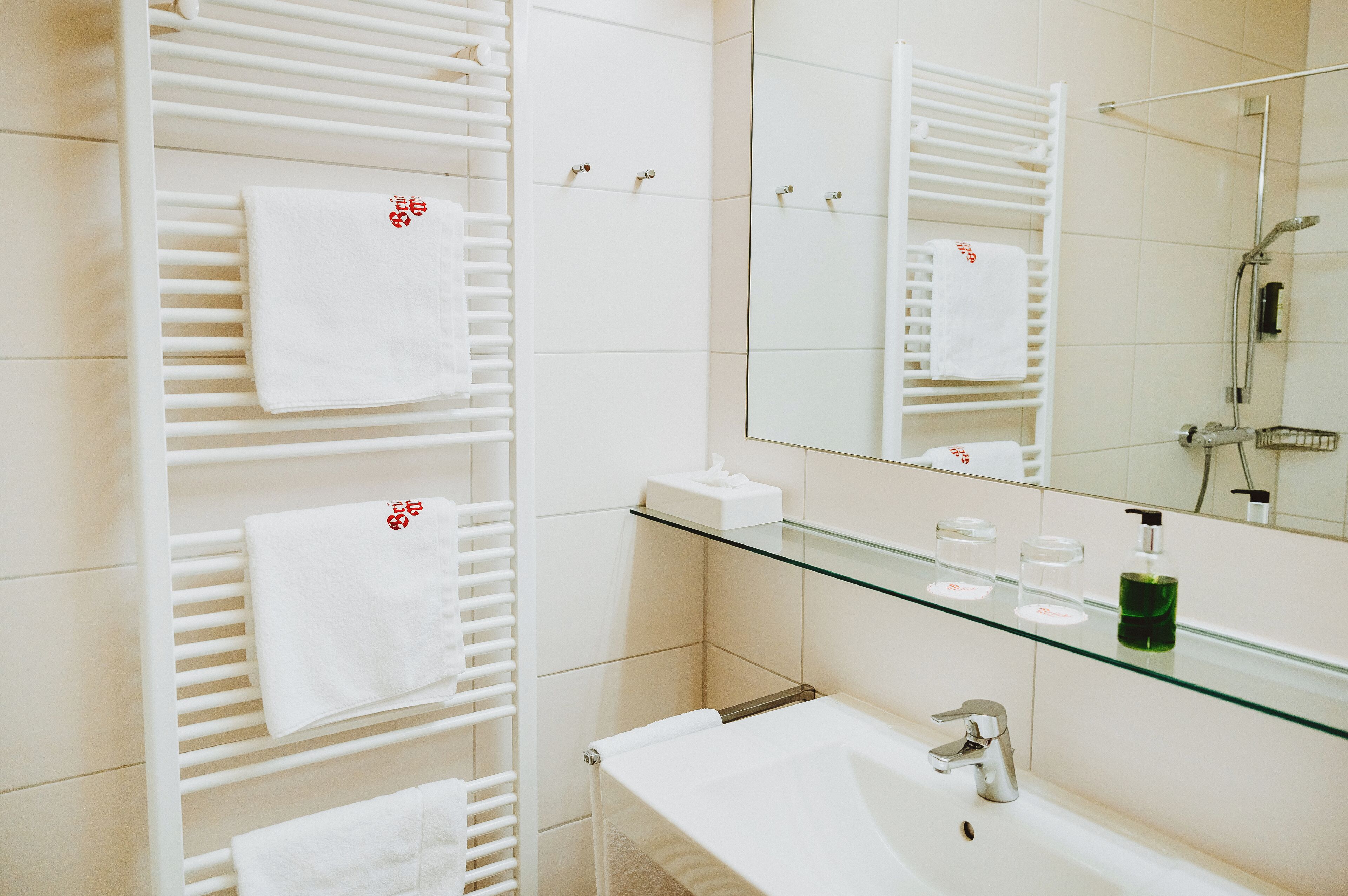 A modern bathroom with white tiled walls, a glass shelf with toiletries, a white sink, and a large mirror.