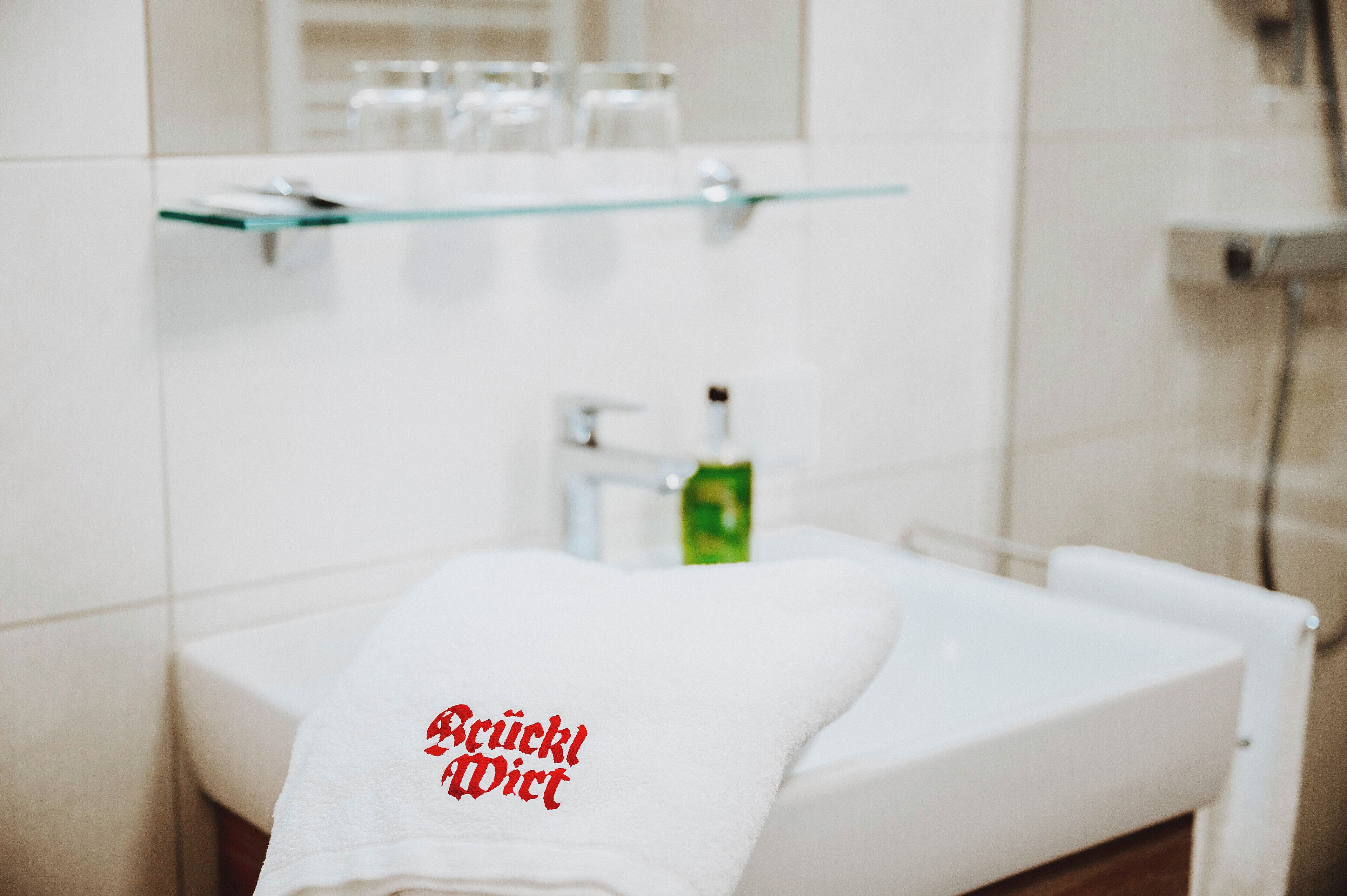 Un baño con un lavamanos blanco, un estante de vidrio arriba y una toalla con texto rojo.