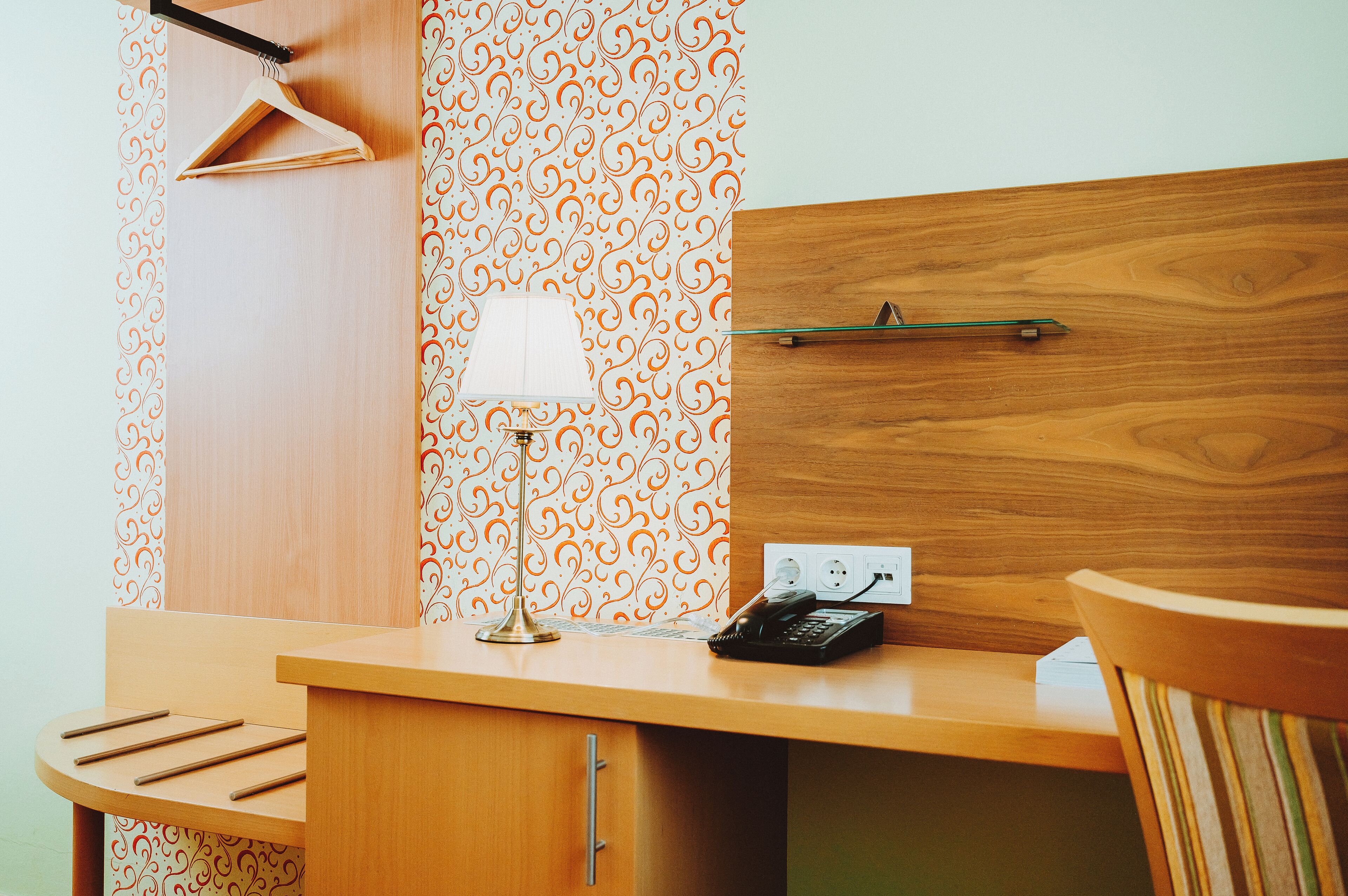 A hotel room with a wooden desk, a lamp, a telephone, and a wall with a patterned wallpaper.