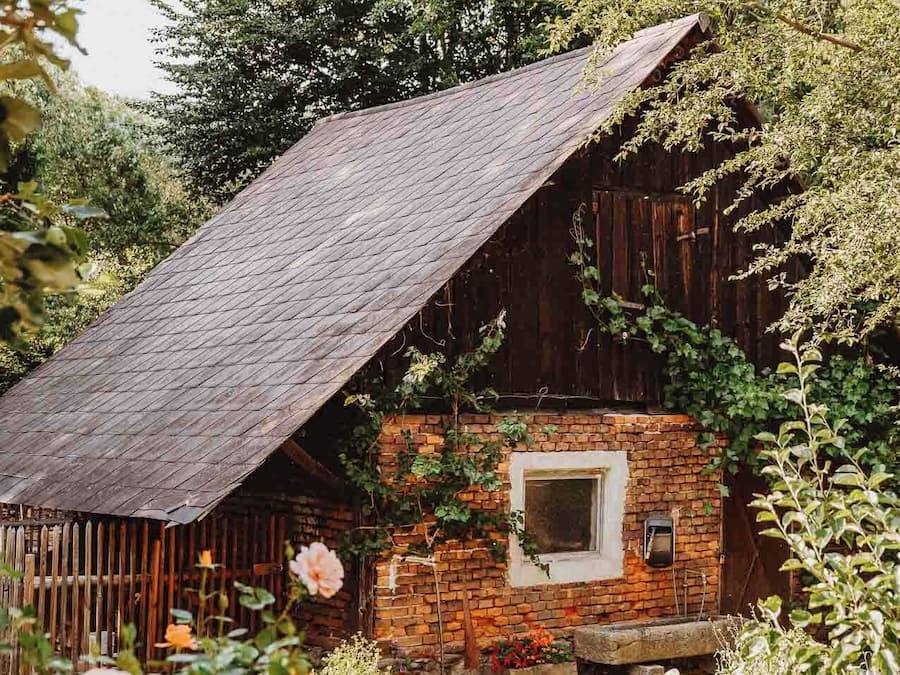 A rustic wooden barn with a sloped roof, surrounded by lush greenery and flowering plants.