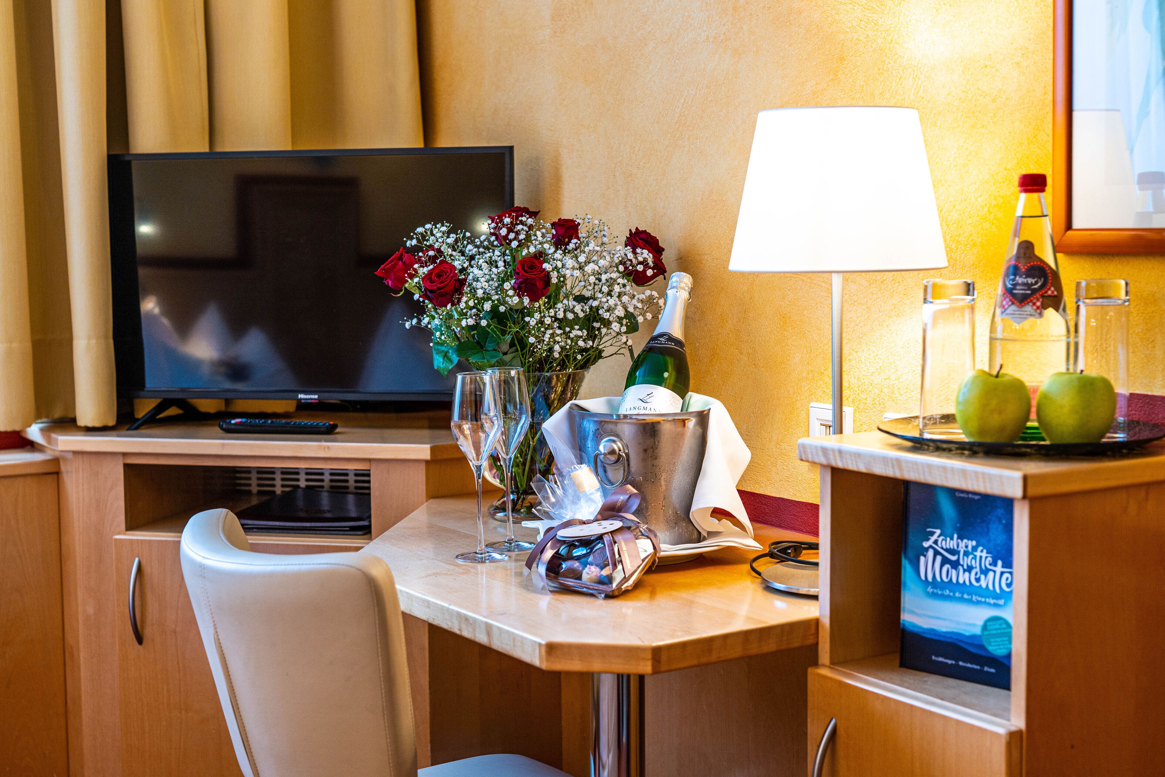 Habitación de hotel con televisor, escritorio con lámpara, florero con flores, botella de champán y menú.