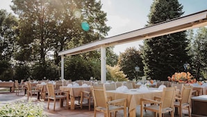 Ein Essbereich im Freien mit runden Tischen und Stühlen, der von einer Pergola bedeckt und von Bäumen und Pflanzen umgeben ist.