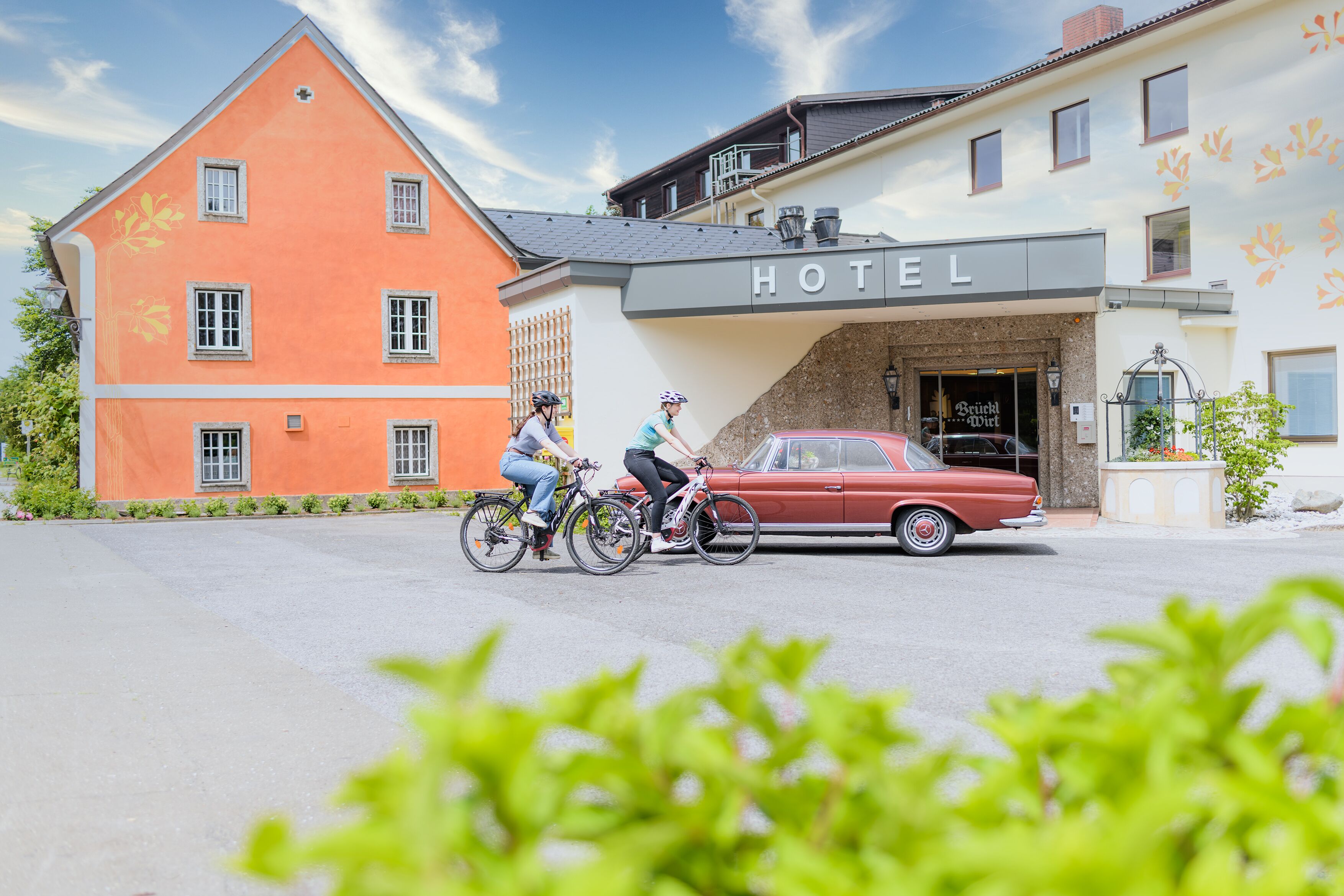 Un hôtel avec une enseigne «HÔTEL» bien visible, une voiture rouge d’époque garée devant, et deux cyclistes qui passent.