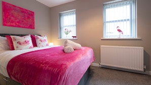 A bedroom with a bed, pink bedding, a radiator, and two windows with curtains.
