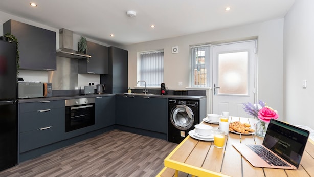 A modern kitchen with dark cabinetry, a built-in oven, a washing machine, and a wooden table with a laptop and breakfast items.