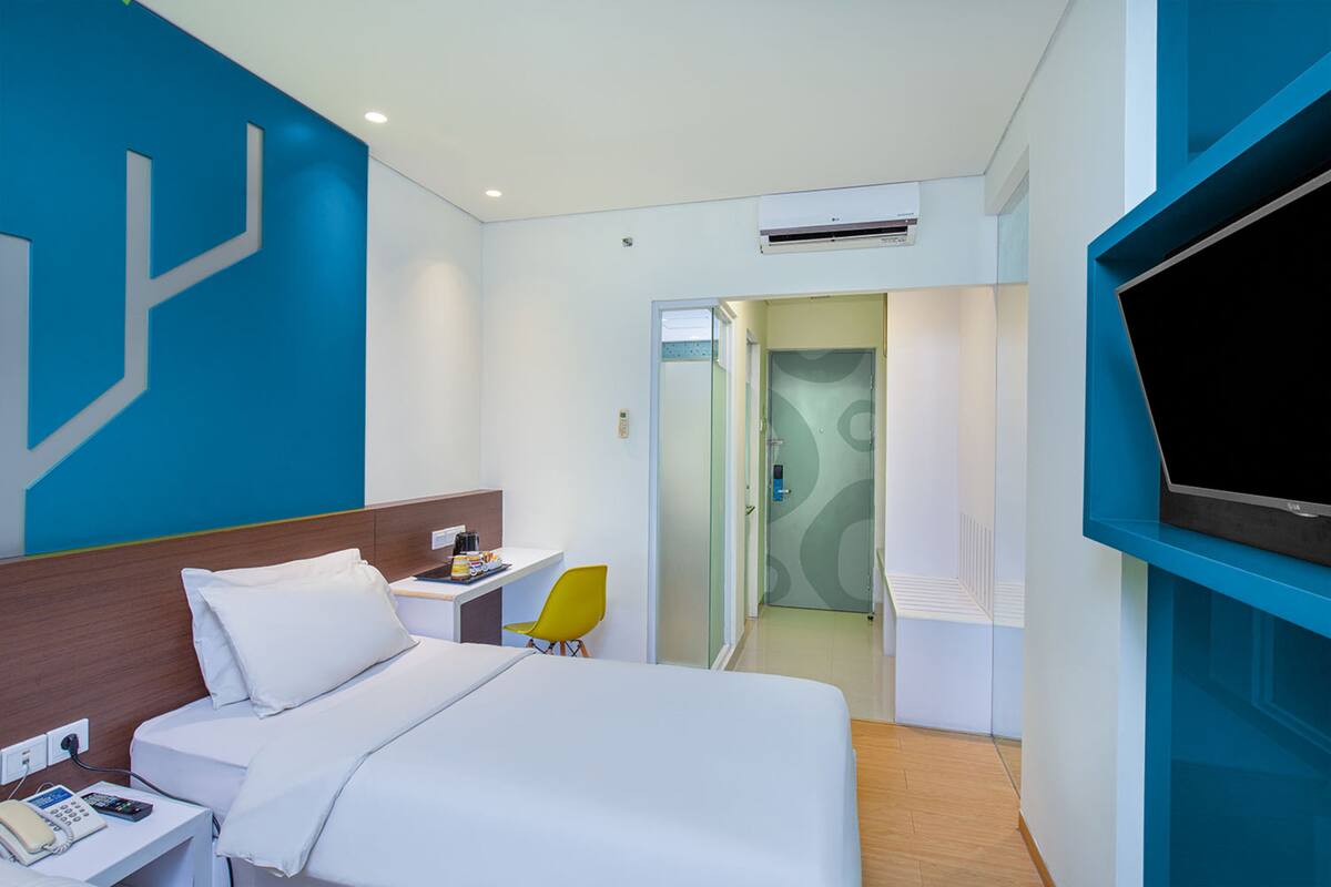 A hotel room with a bed, a desk, a chair, a TV, and an air conditioning unit.