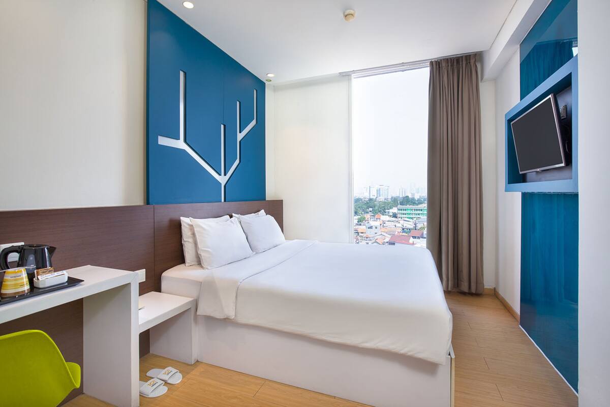 A hotel room with a bed, a desk with a coffee maker, a TV, and a window with a city view.