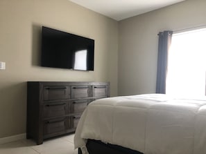Une chambre à coucher comprenant un lit, une commode, un téléviseur à écran plat fixé au mur et une fenêtre avec des rideaux.