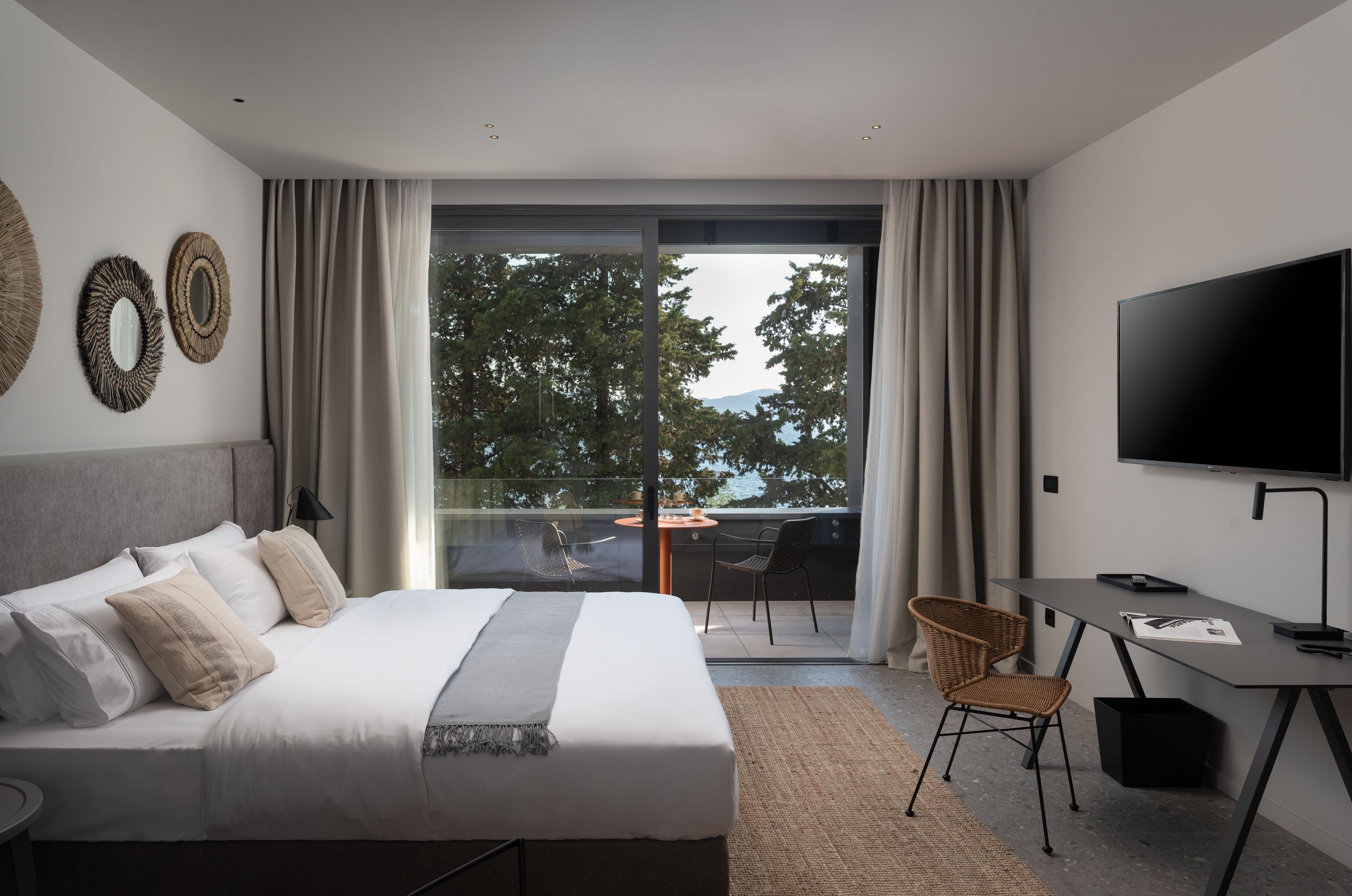 A modern hotel room with a large bed, a desk, a TV, and a balcony with a view of trees and mountains.