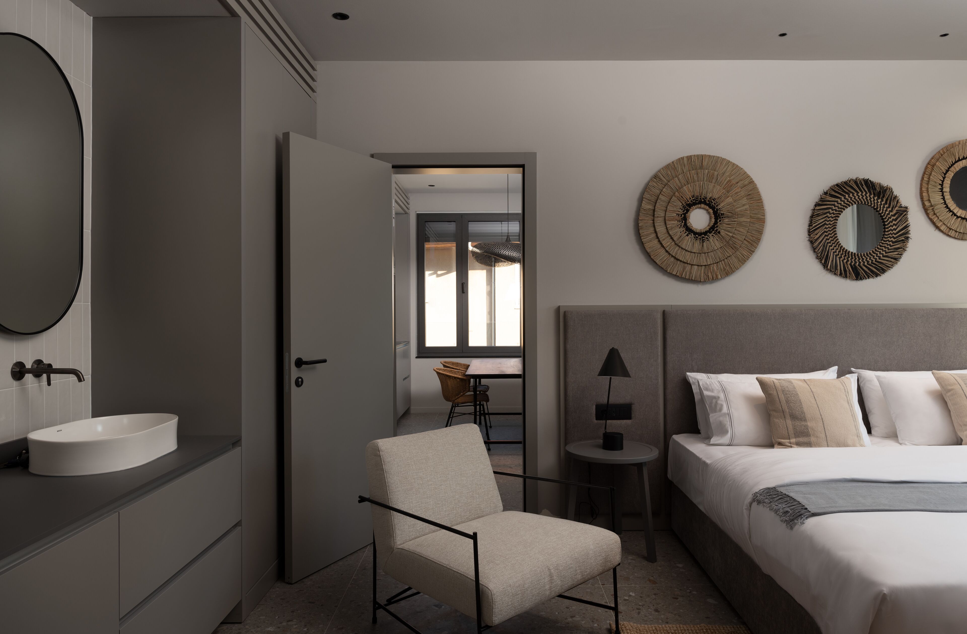 Un dormitorio moderno con una cama grande, una mesita de noche, una silla, un espejo redondo y un baño con lavabo e inodoro.