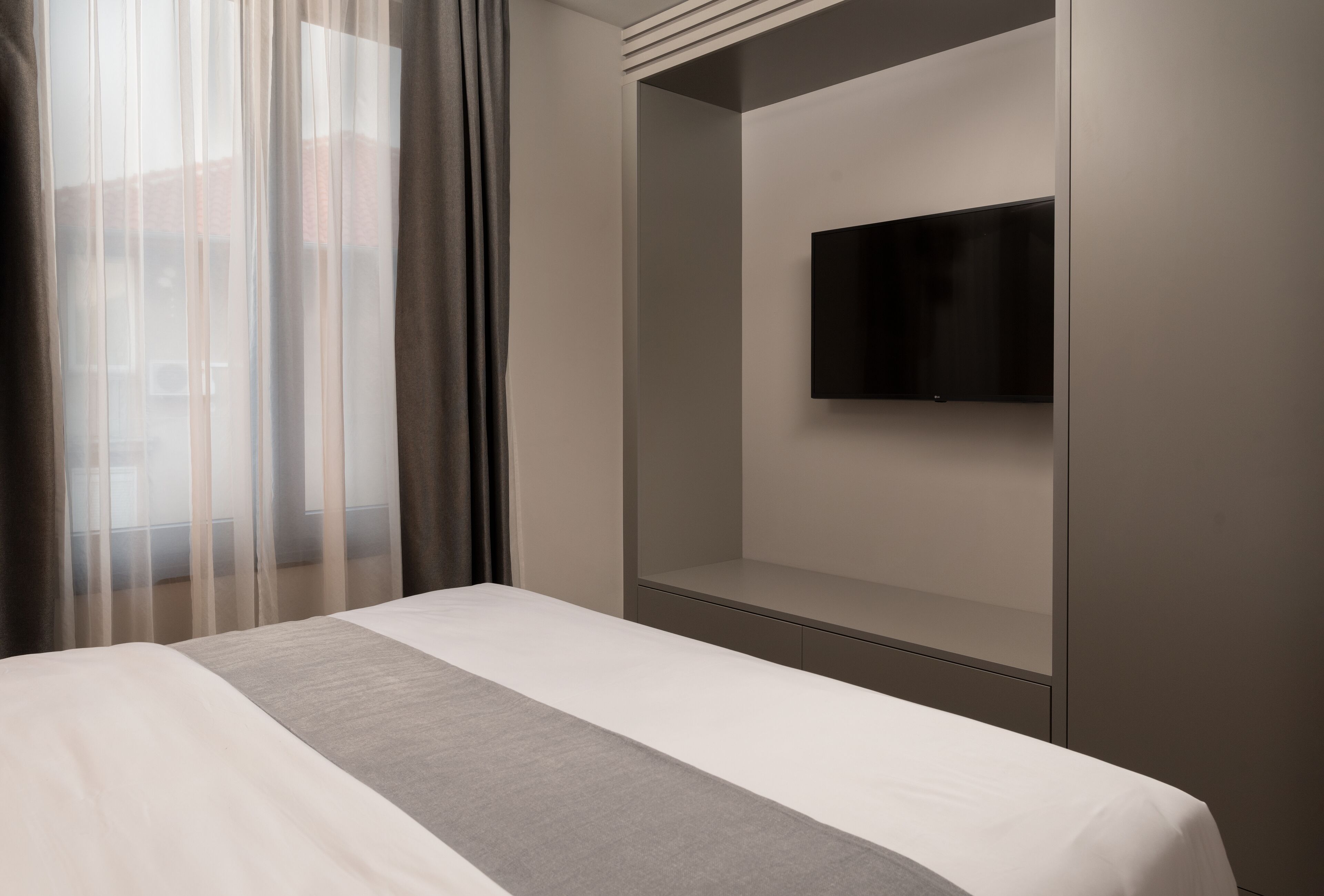 Habitació d'hotel amb un llit, una televisió penjada a la paret i una finestra amb cortines transparents.