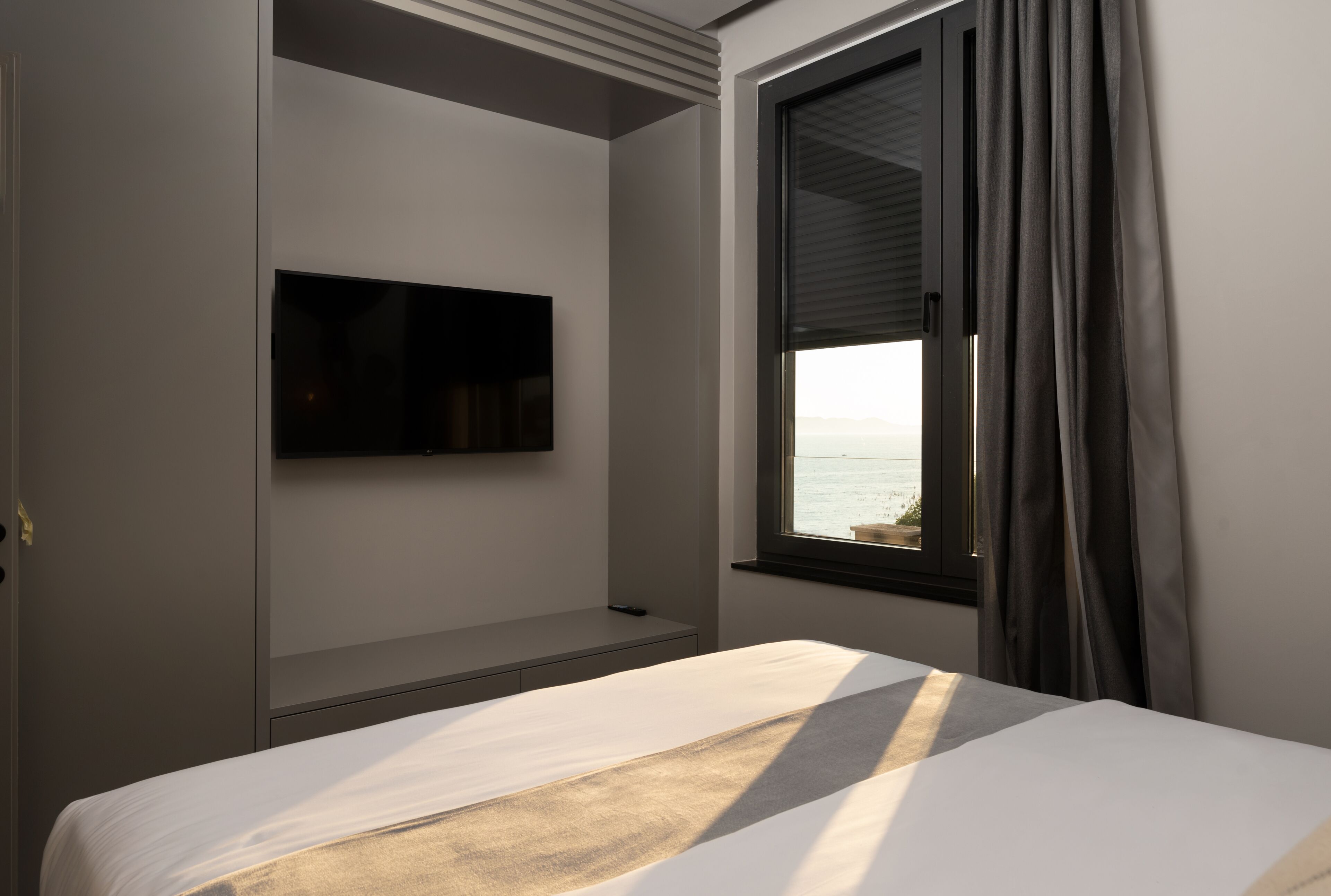 A hotel room with a bed, a mounted TV, a window with curtains, and a view of the sea.