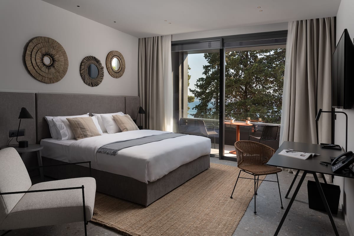 A modern bedroom with a large bed, a desk, and a chair. There are two wall mirrors and a window with a view of trees.