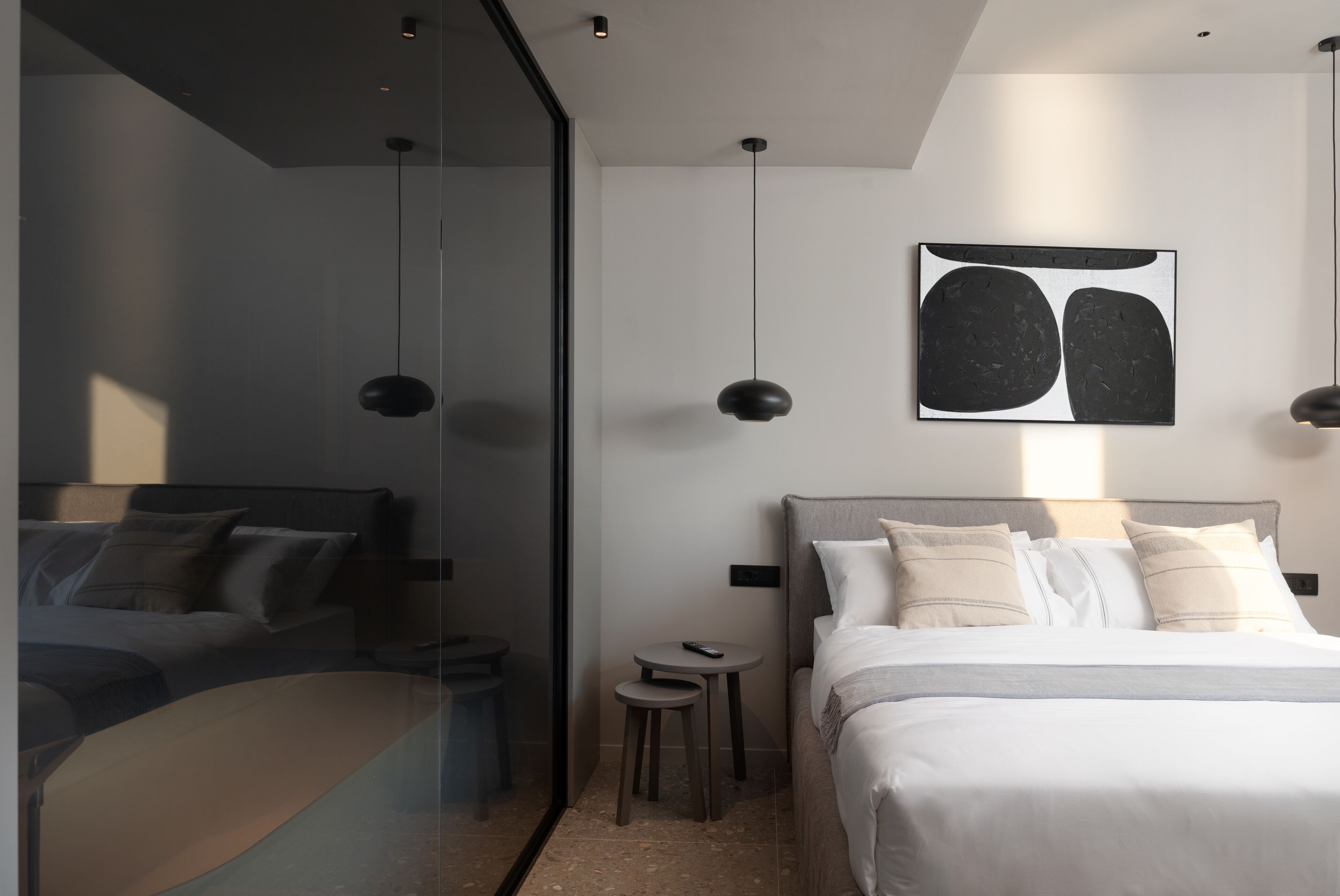 A modern hotel room with a glass partition, a bed with white bedding, a small round table, and a pendant light.