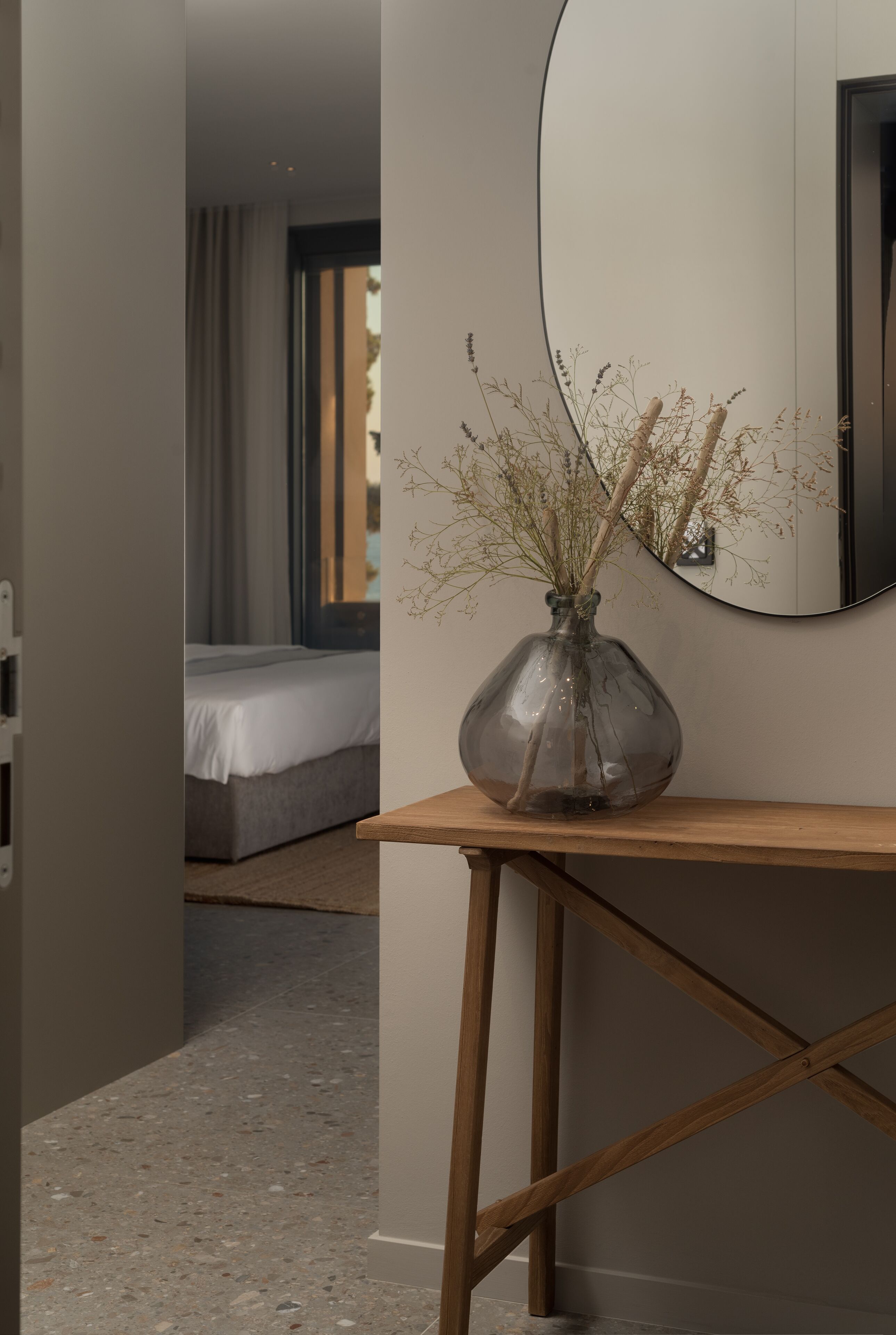 Une chambre d’hôtel avec un lit, une table de chevet en bois ornée d’un vase contenant des plantes séchées, et un miroir rond accroché au mur.