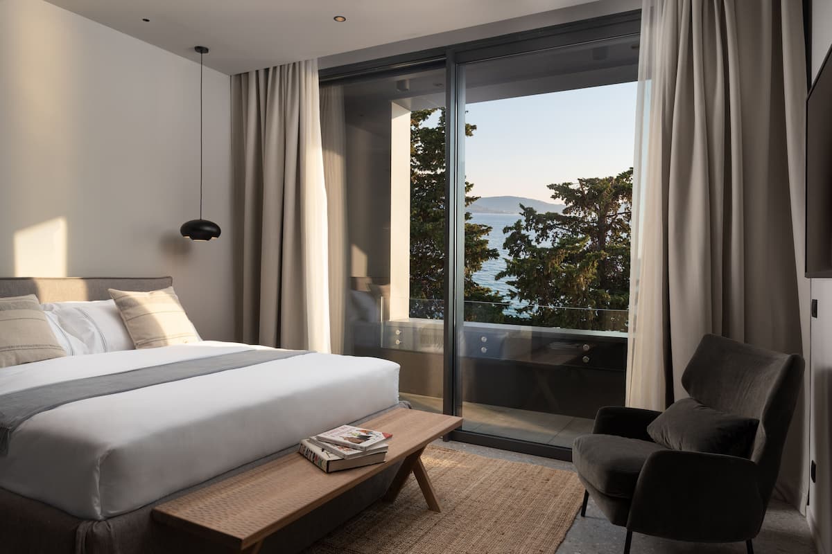 A modern hotel room with a large bed, a reading bench, a chair, and a balcony with a view of the sea.