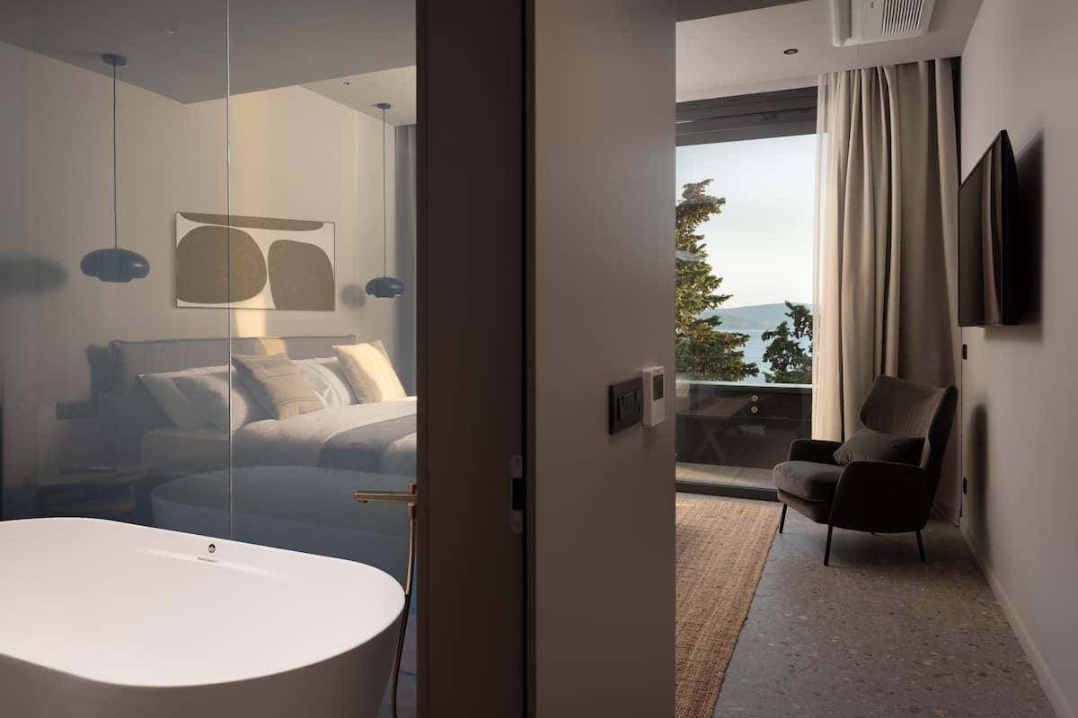 A modern bedroom with a large bed, a white bathtub, a chair, and a TV on the wall.