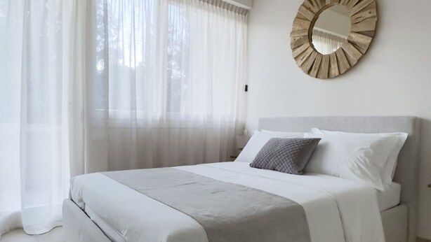 A modern bedroom with a large bed, sheer curtains, a round mirror, and a tiled floor.