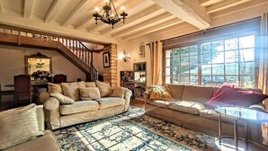 A cozy living room with two sofas, a patterned rug, a chandelier, and a large window with a view of the outdoors. - La Maison bleue (Cuncy-lès-Varzy)