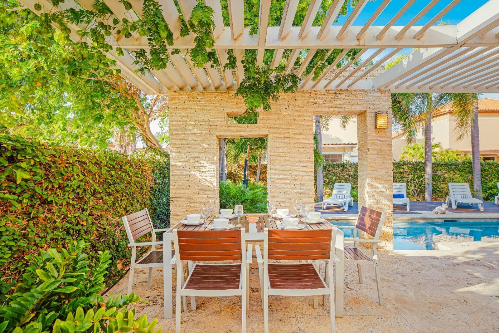 Un espace extérieur aménagé pour les repas, avec une pergola, des murs en briques et une piscine en arrière-plan.