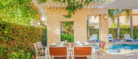 Un espace extérieur aménagé pour les repas, avec une pergola, des murs en briques et une piscine en arrière-plan.