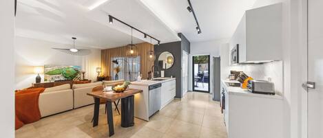 Eine moderne Küche mit einer zentralen Kochinsel, ein Essbereich mit einem Holztisch und ein Wohnzimmer mit Sofa und Gemälde.