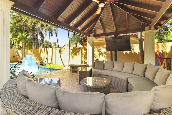 A covered outdoor seating area with a pool, wicker furniture, and a flat-screen TV. - Bocobay Gold Coast Resort (Noord)
