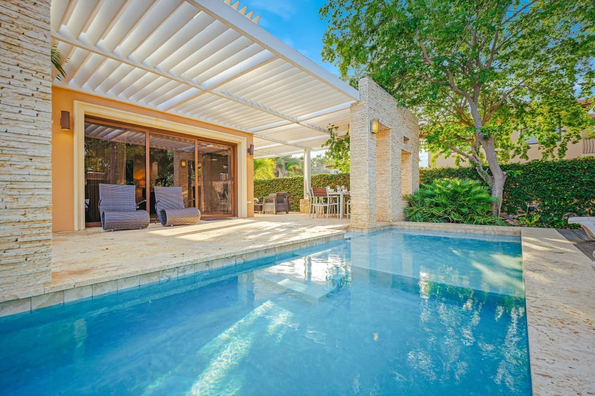 Ein Schwimmbecken mit überdachtem Patio, einer Steinmauer und Gartenmöbeln.
