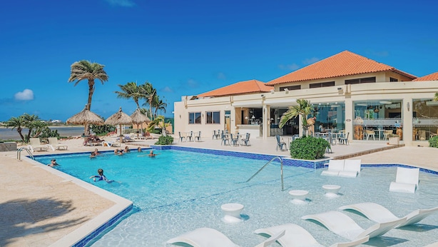 Une piscine avec des chaises longues, des palmiers et un bâtiment doté d’un toit en tuiles.
