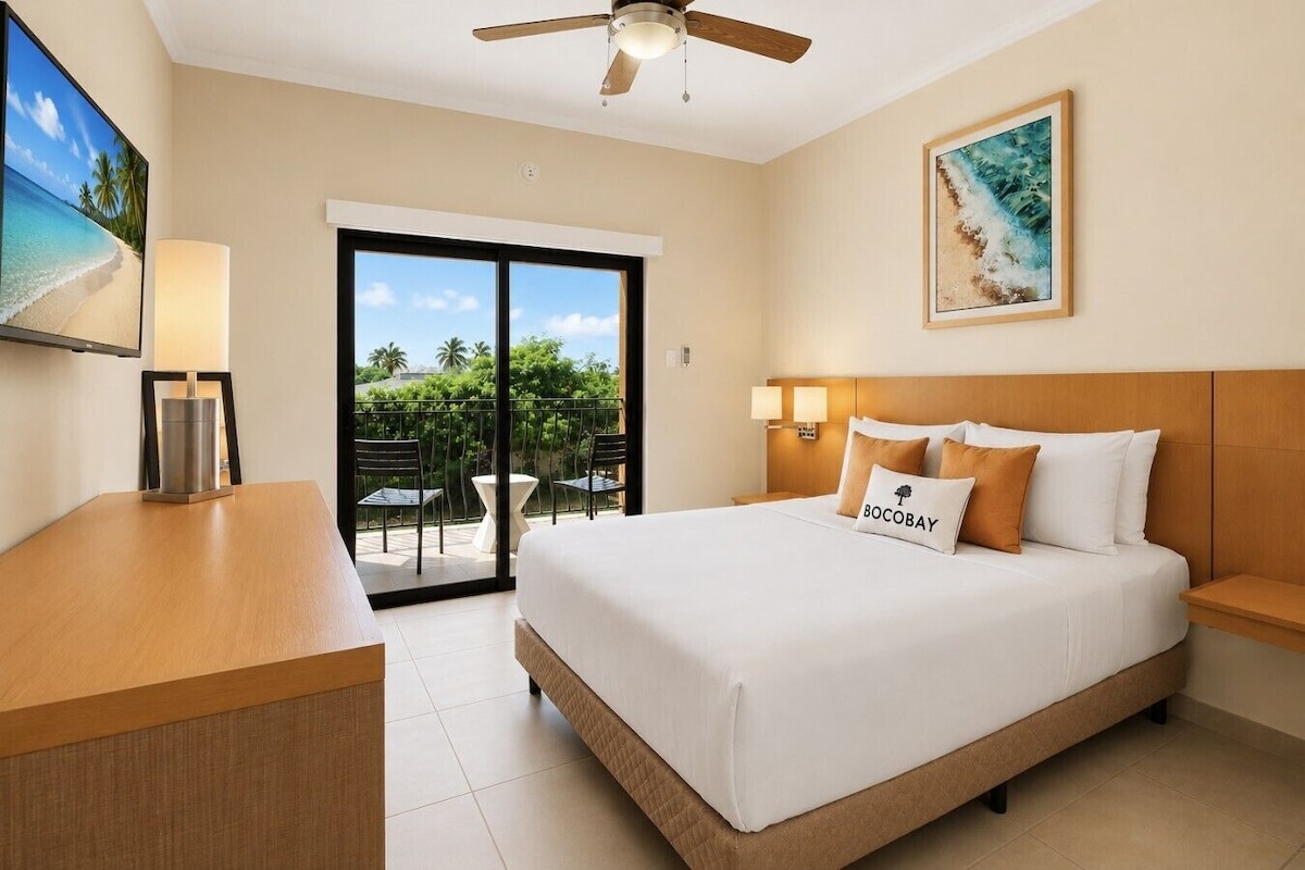 Habitación de hotel con cama, escritorio, silla, ventilador de techo, un cuadro y vistas al exterior.