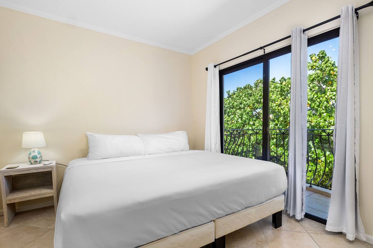 Ein Schlafzimmer mit einem Bett, einem Nachttisch, einer Lampe und Blick auf Grün hinter einem Fenster.
