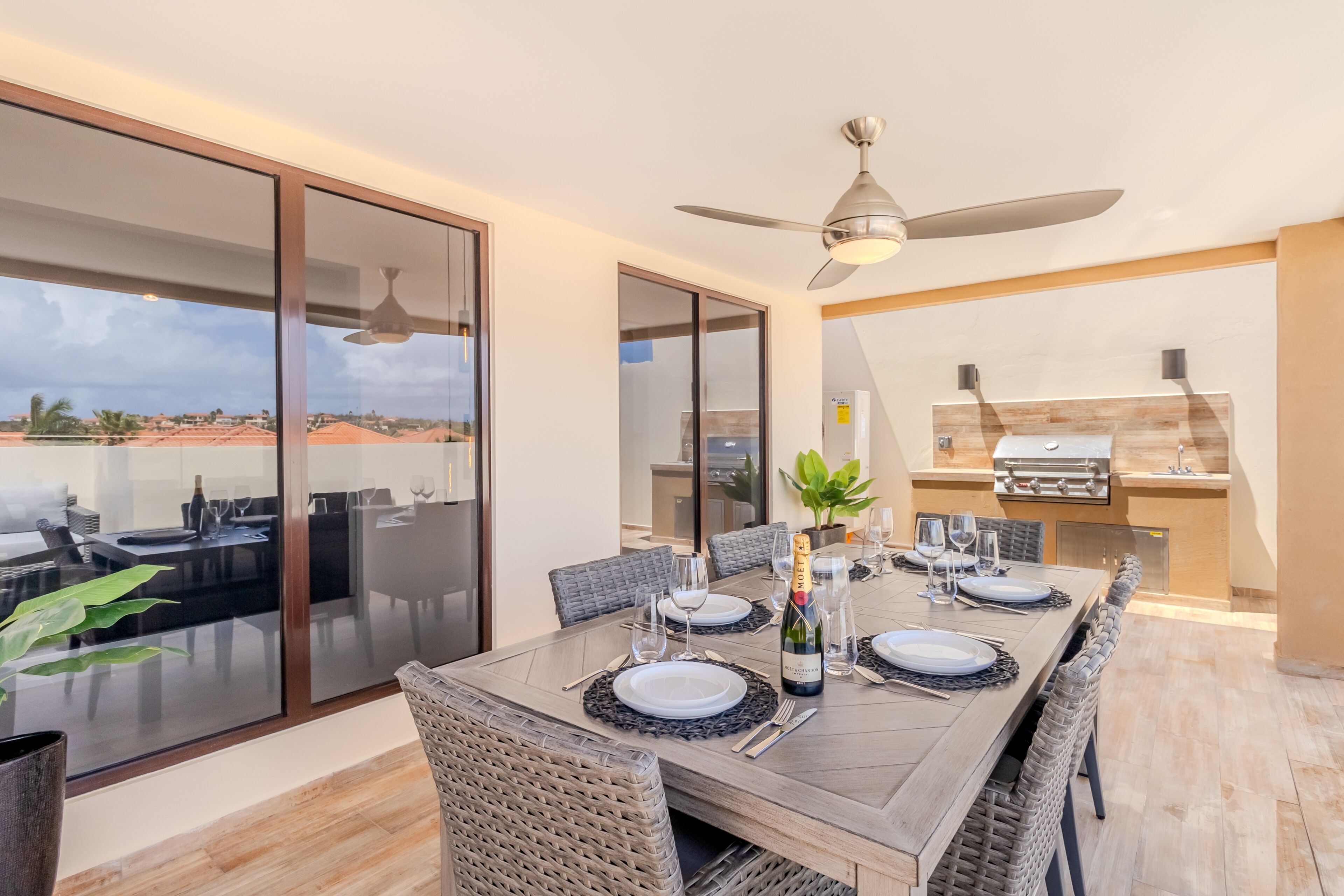 Un comedor con una mesa grande dispuesta para seis comensales, un ventilador de techo y una zona de cocina con una parrilla.