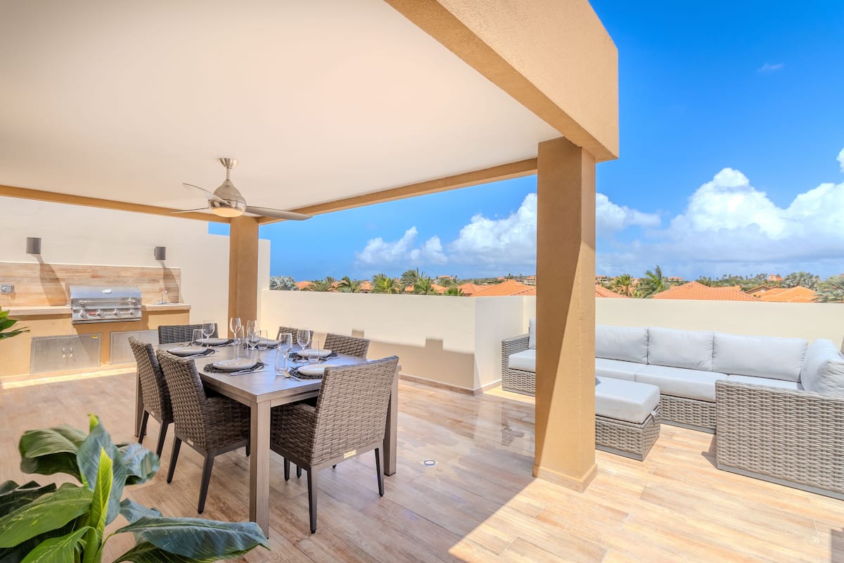 Eine Außenpatio mit einem Esstisch, gedeckt für vier Personen, umgeben von Korbmöbeln, einem Grill und Blick auf die Landschaft.