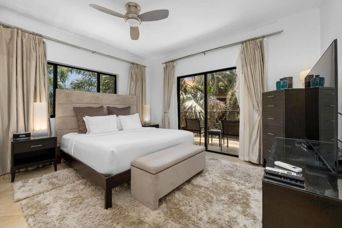 Ein modernes Schlafzimmer mit einem großen Bett, einer Bank, einem Fernseher und einem Balkon mit Vorhängen.