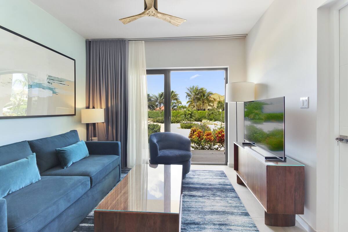 Ein modernes Hotelzimmer mit Sofa, Couchtisch, Fernseher und Blick auf Palmen und einen Garten.