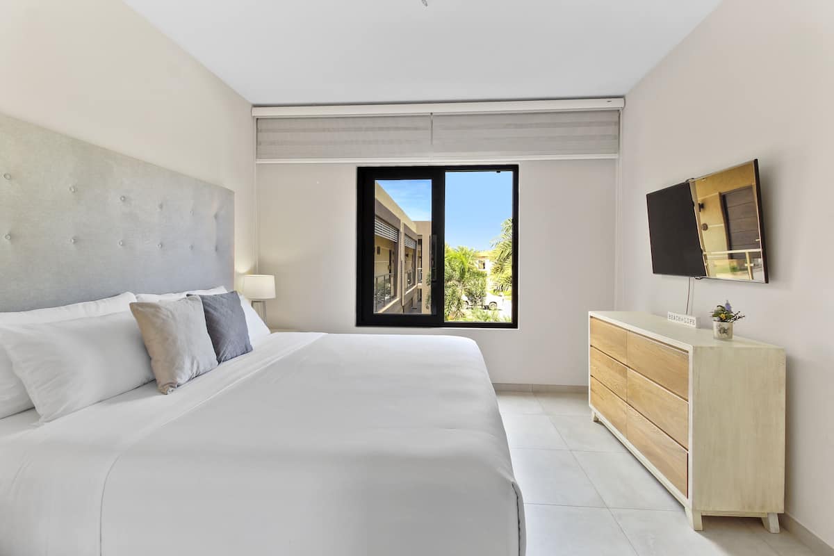 Ein Schlafzimmer mit einem großen Bett, einer Holzkommode, einem Flachbildfernseher und einem Fenster mit Blick auf Gebäude und Grünflächen.