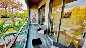 Un balcón con barandilla de vidrio, mobiliario exterior y vistas a un jardín y árboles.