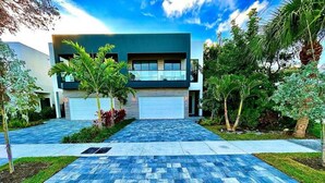 Una casa moderna de dos pisos con garaje, rodeada de abundante vegetación y un camino de entrada con baldosas azules estampadas.