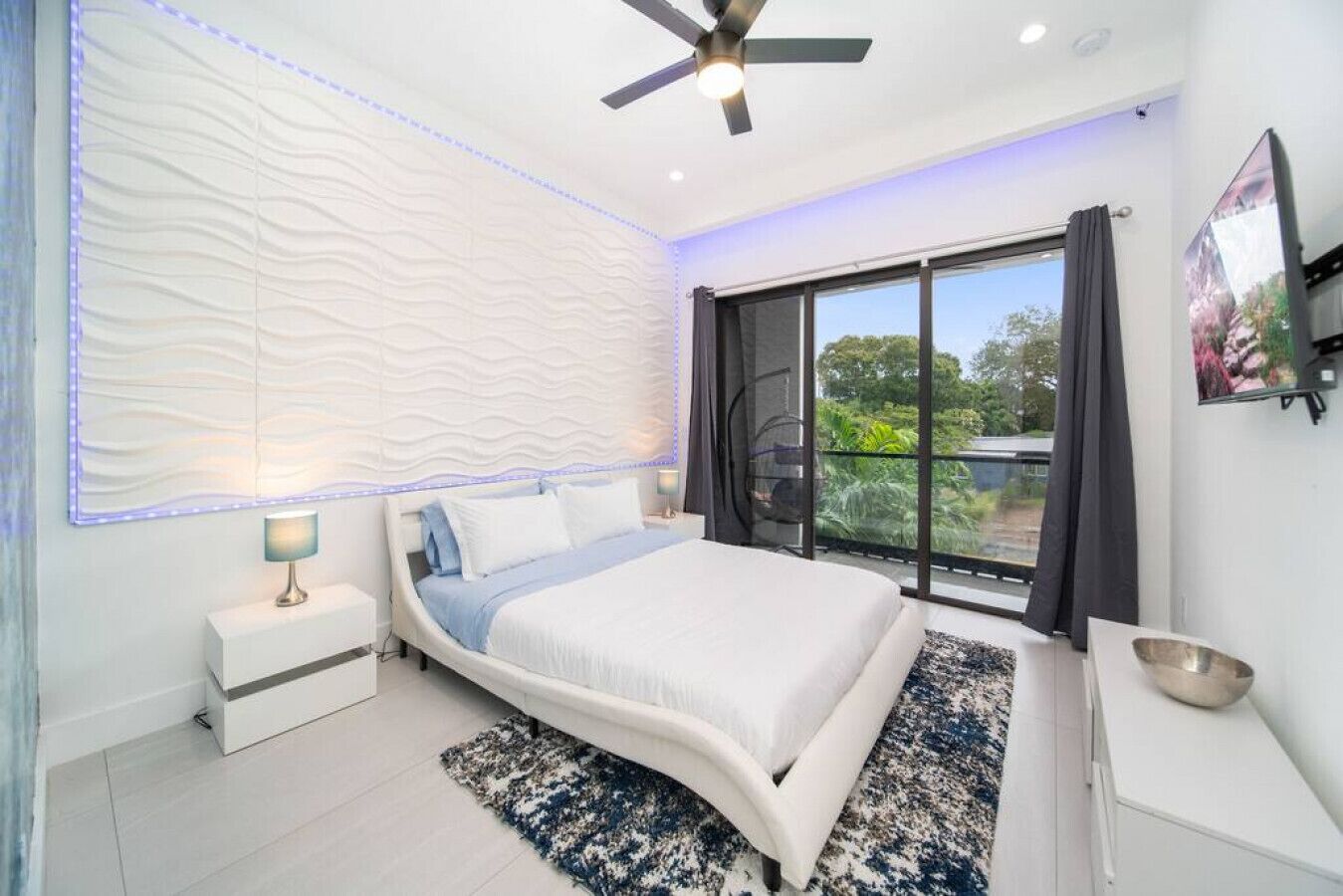 A modern bedroom with a curved bed, a ceiling fan, a large window with curtains, and a nightstand with a lamp.