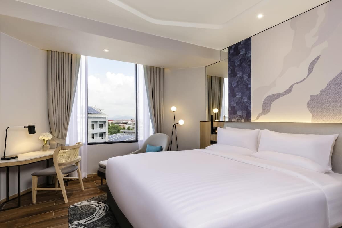 A modern hotel room with a large bed, a desk with a chair, a standing lamp, and a view of the cityscape through the window.