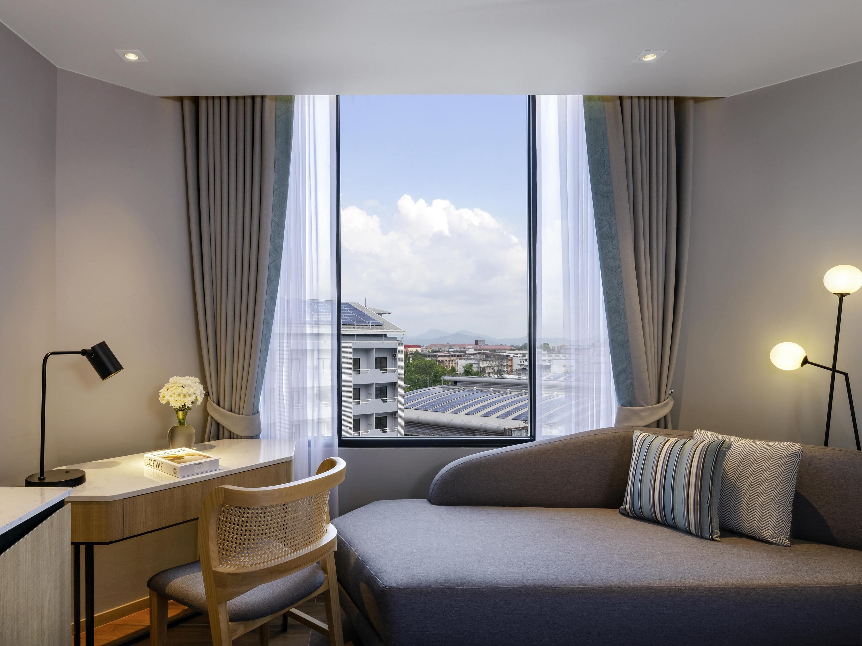 Una habitación de hotel moderna con escritorio, silla y sofá, ventanas amplias con cortinas, y vista a edificios y paneles solares.