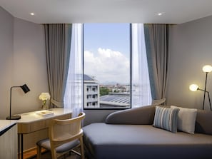 A modern hotel room with a desk, chair, and sofa, large windows with curtains, and a view of buildings and solar panels. - Novotel Rayong Star Convention Centre (Rayong)