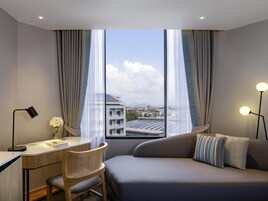 Una habitación de hotel moderna con escritorio, silla y sofá, ventanas amplias con cortinas, y vista a edificios y paneles solares.